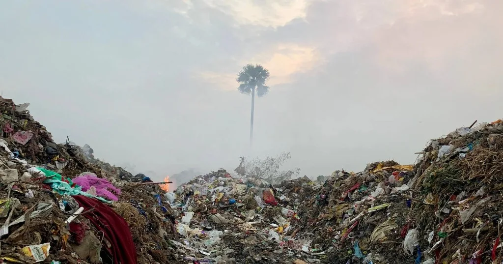 Open dump of plastic waste in Kerala, with smoke rising around a palm tree